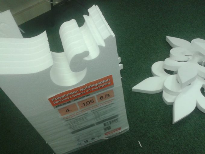 Several identical scroll profiles cut straight through a full pack of EPS polystyrene insulation board, with a cut foam flower beside it