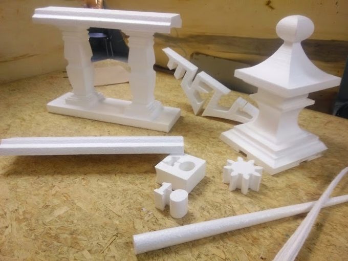 A batch of white EPS foam parts hot-wire cut on a FoamCube — a two-column balustrade, a finial, a Greek-key fret, gears and cylinders — on a workbench
