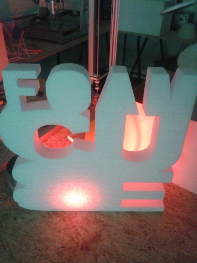 Flat 2-axis hot-wire-cut foam lettering, backlit with warm light, on the machine bed